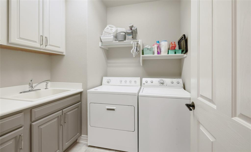 Laundry room includes upper and lower cabinets, racks and a useful wash basin with luxury vinyl tile underfoot.