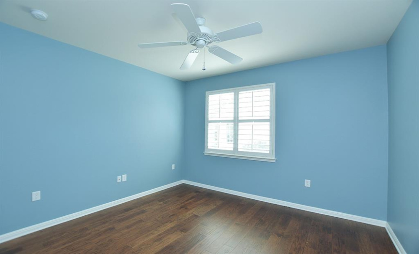 The guest bedroom with wood floors has a shuttered window looking to the side of the home.
