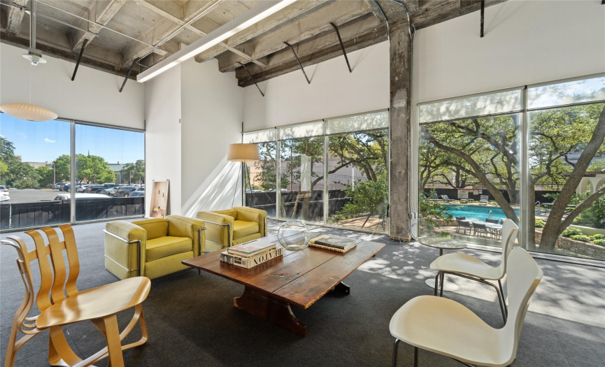 In this spacious office living area, natural light floods the room through expansive floor-to-ceiling windows. The lush greenery of the outdoor pool area is visible through the glass, creating a serene and inviting atmosphere.