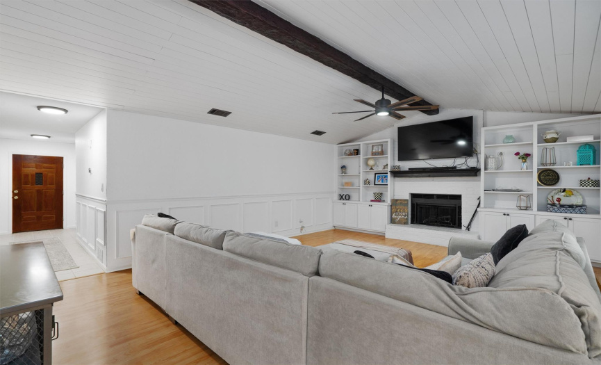 The renovated living room provides timeless elegance with vaulted ceilings wrapped in shiplap, elegant wainscoting, a wood-burning fireplace, custom painted mantelpiece, custom built-ins, and vinyl plank flooring. 