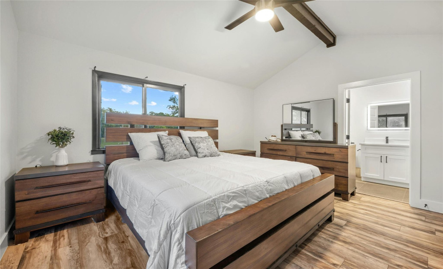 The primary bedroom is located in the back of the house and has an incredible view of the backyard. The room has a vaulted ceiling with a wood beam, a ceiling fan, and beautiful wood-looking vinyl floors.