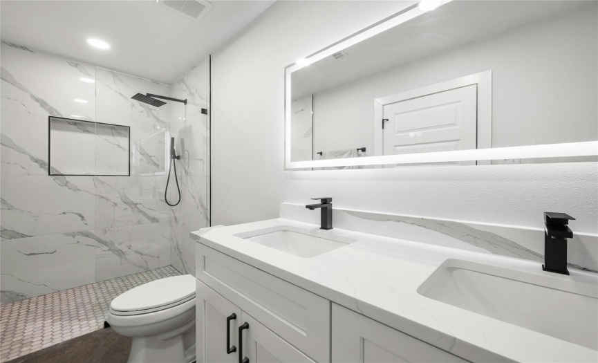 The modern owner's ensuite bathroom has a double vanity with a beautiful quartz countertop with rectangular undermount sinks, a large frameless LED mirror, black cabinet pulls, elegant tile flooring and a large walk-in shower with a custom niche, rain showerhead and shower wand.