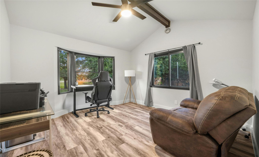 The large dedicated office has a soaring vaulted ceiling with a wood beam, wood-looking vinyl flooring, modern ceiling fan, and many windows that provide natural lighting with a view of your manicured front yard.
