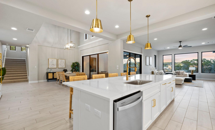 The warm glow of brass pendant lighting suspended above the expansive kitchen island creates an inviting space that is seamlessly connected to both the dining and living areas.