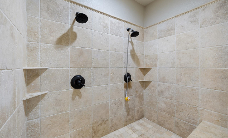 The gorgeous oversized walk-in shower is surrounded in tile and features two side by side shower heads and bench seating. 