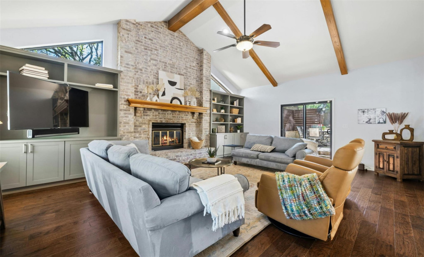 The remarkable living room is bathed in natural light, accentuating its vaulted wood-beamed ceiling, floor-to-ceiling stone fireplace, and custom built-ins