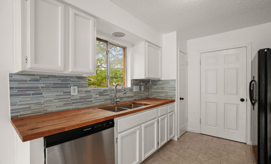 Galley type kitchen has been updated with butcher block counters, glass tile backsplash, painted cabinets and tile flooring. 