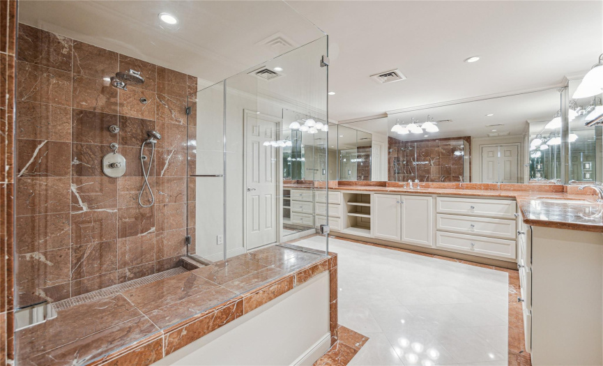 The primary bathroom is just behind french doors. It features a glass shower with bench, double sinks, water closet, and storage cabinetry and drawers.